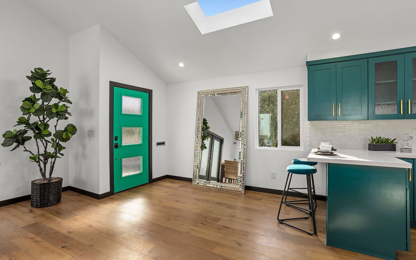 Interior with teal door, cabinets, and island. Hardwood floors, large mirror, and potted tree.