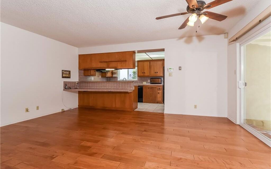 Empty living room with wooden floors, kitchen bar, and sliding glass door.