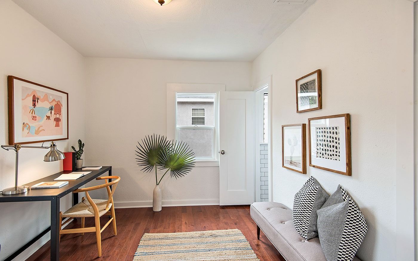 Small home office with desk, artwork, bench, and a faux tree. White walls, wood floors.