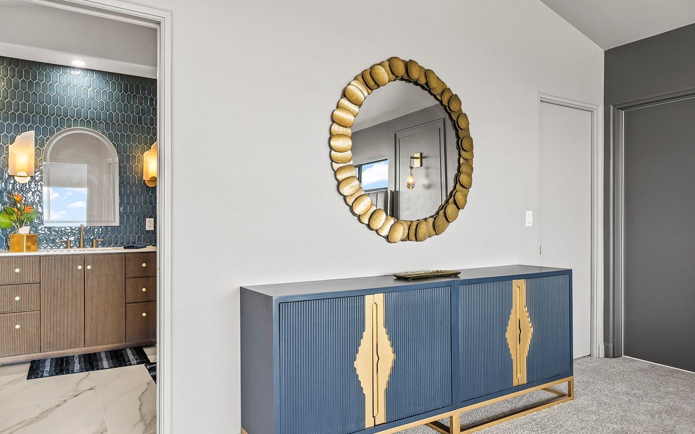 A blue cabinet with gold accents and a decorative mirror on a white wall, with a bathroom visible on the left.