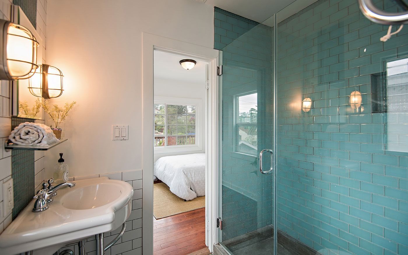 Bathroom with a glass shower, teal tile, white sink, and an open door to a bedroom with a white bed.
