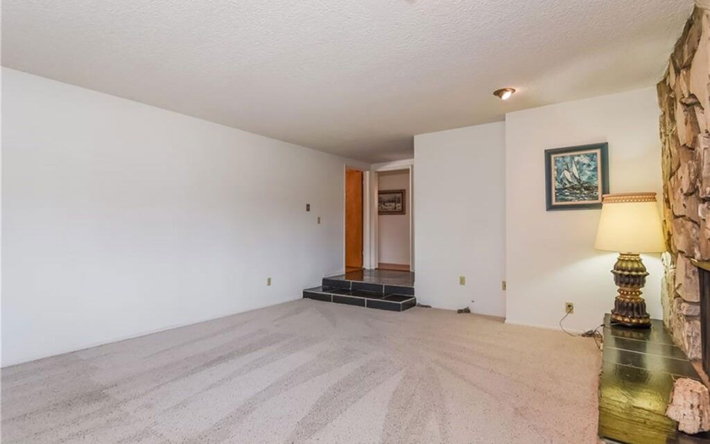 Empty living room with beige carpet, white walls, stone fireplace, and entryway.