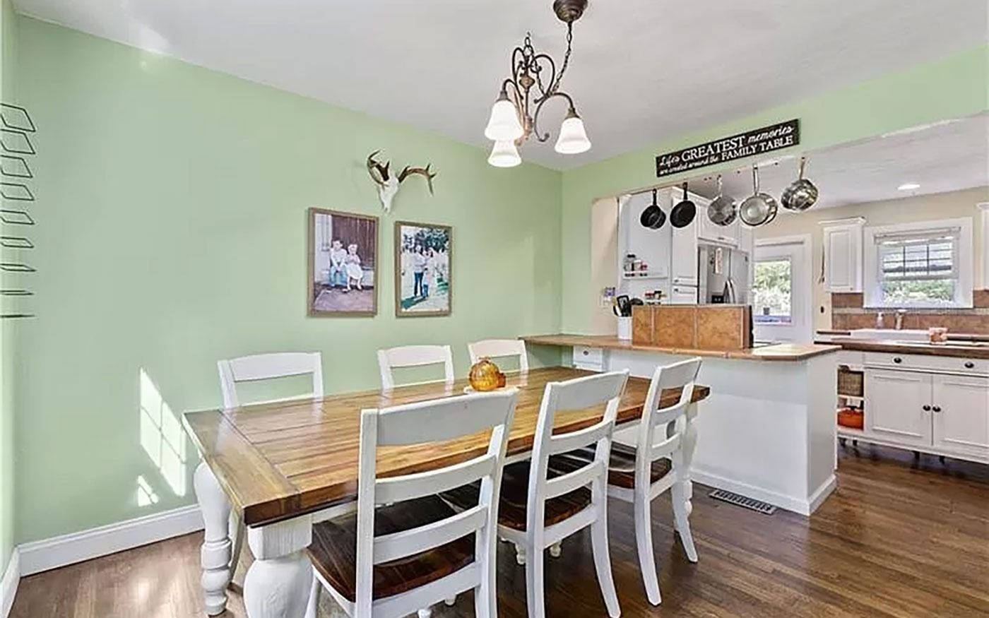 Dining room with a wooden table and white chairs, light green walls, and kitchen in the background.