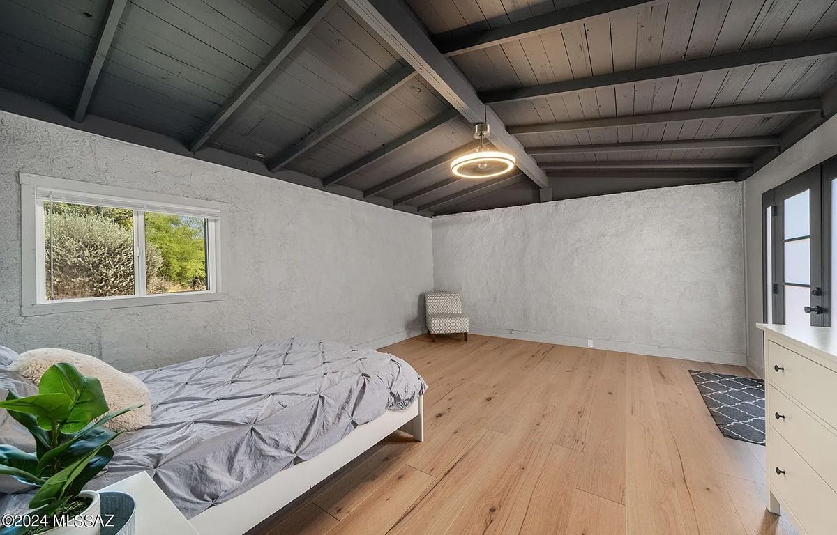 Bedroom with wooden floor, bed, window, armchair, and a doorway, with a dark ceiling.