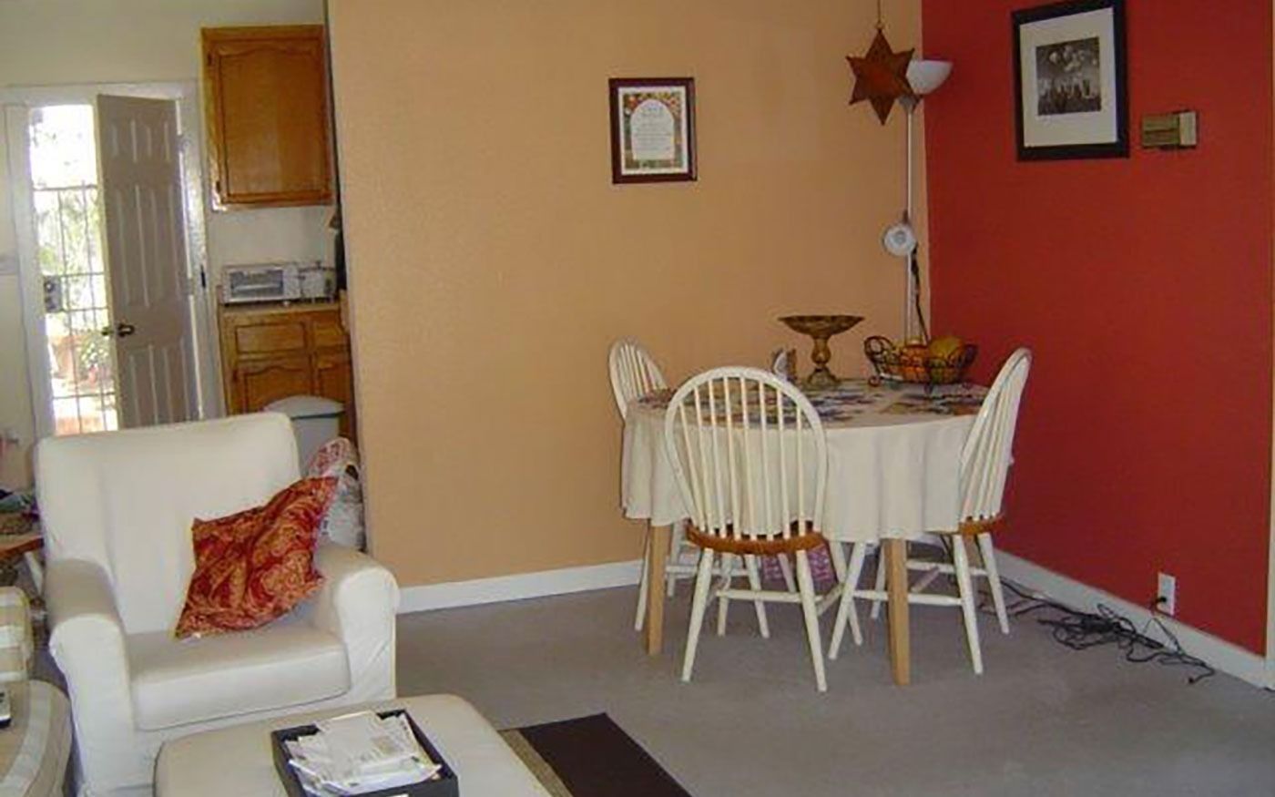 A dining room with an orange and red painted wall, a round table with four white chairs, and a white armchair.