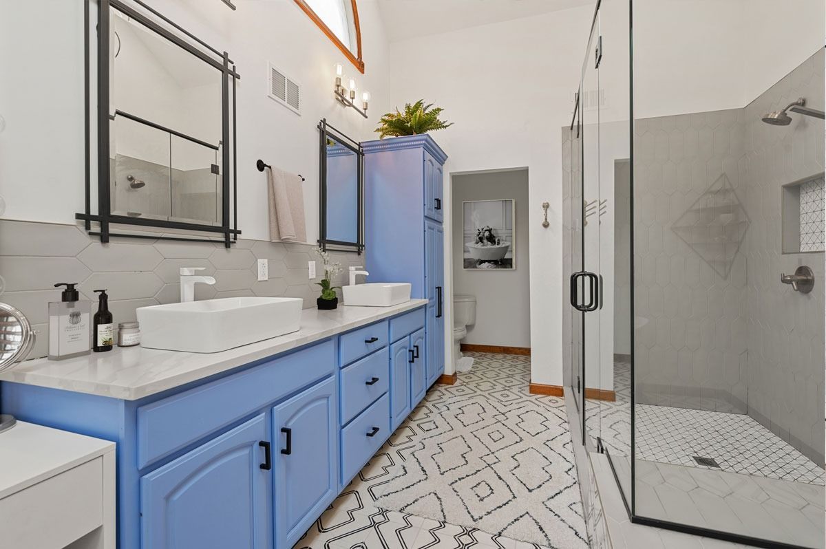 Blue vanity bathroom with two sinks, patterned floor, glass shower, and blue storage cabinet.