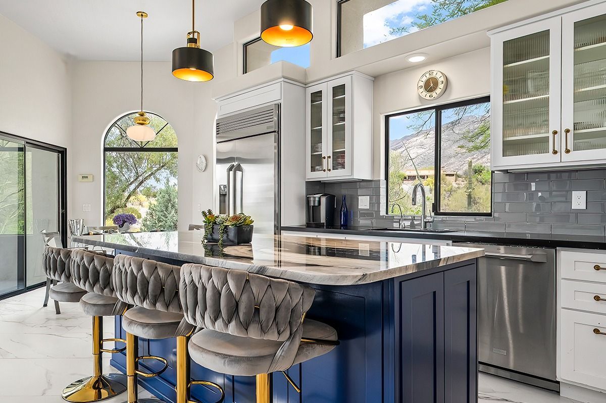 Elegant kitchen with blue island, marble countertops, and gold accents, featuring bar stools and large windows.