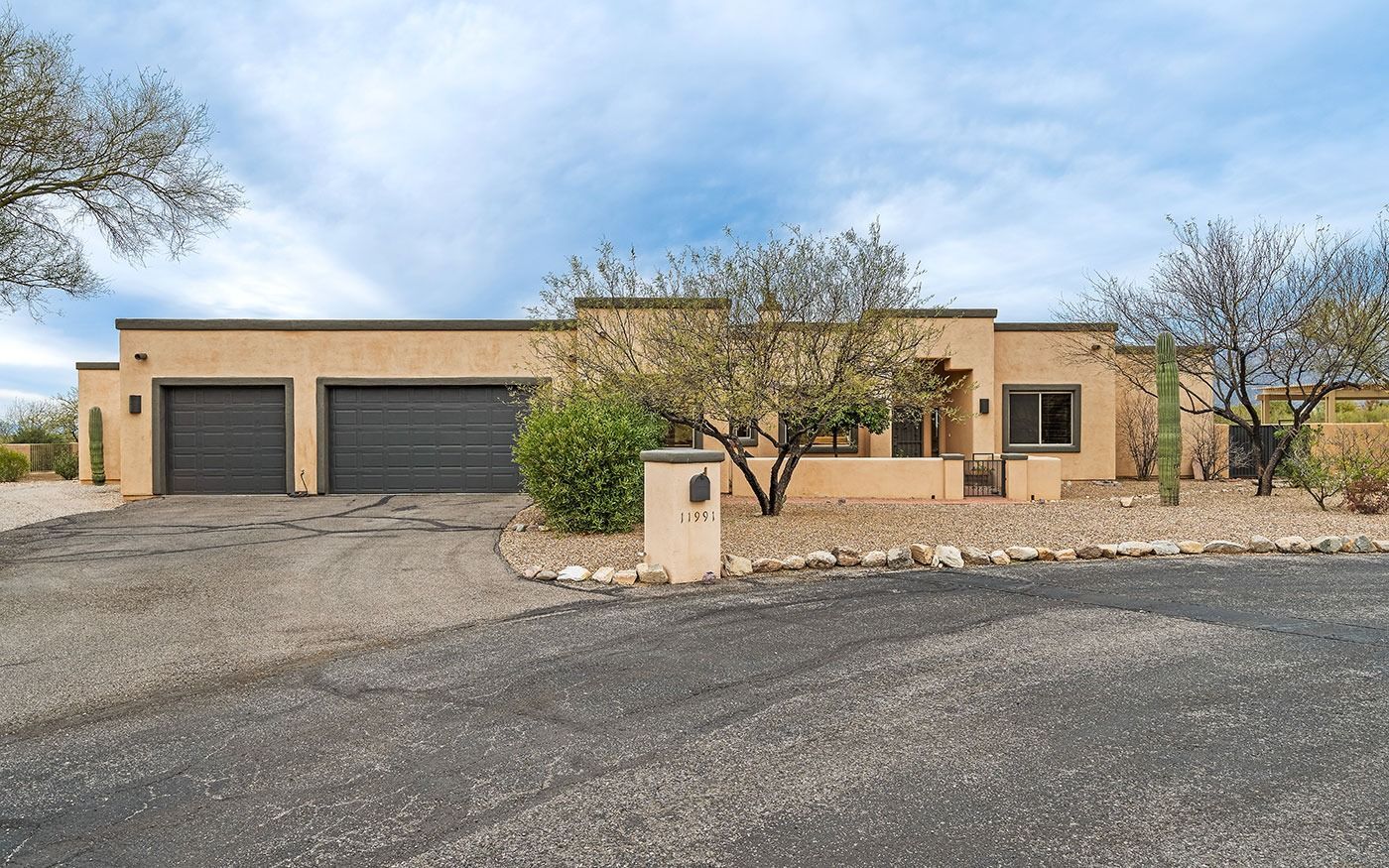 Tan-colored ranch-style house with a gravel driveway and two-car garage, under a cloudy sky.