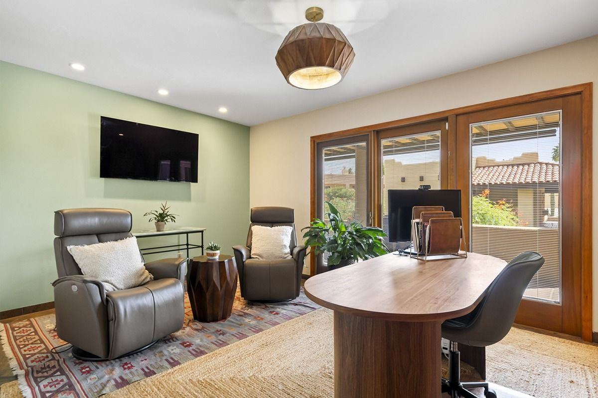 A home office with desk and chairs, two recliners, TV, rug, and large windows with a view.