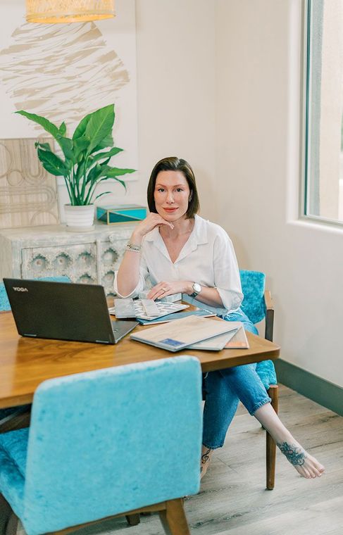 Woman sitting at a table with a laptop, blue chairs, and decorative plant, smiling.