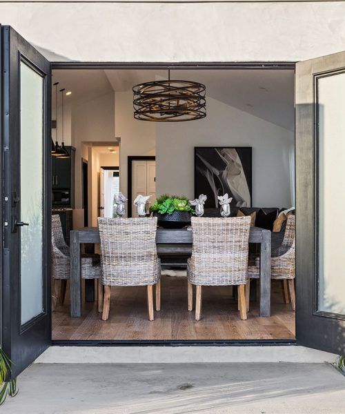 Dining room with open doors. Wooden table with woven chairs, centered by chandelier and art.