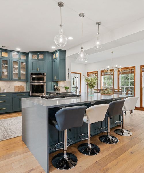 Blue kitchen with an island, light-colored countertops, and pendant lights. Barstools are lined up at the island.