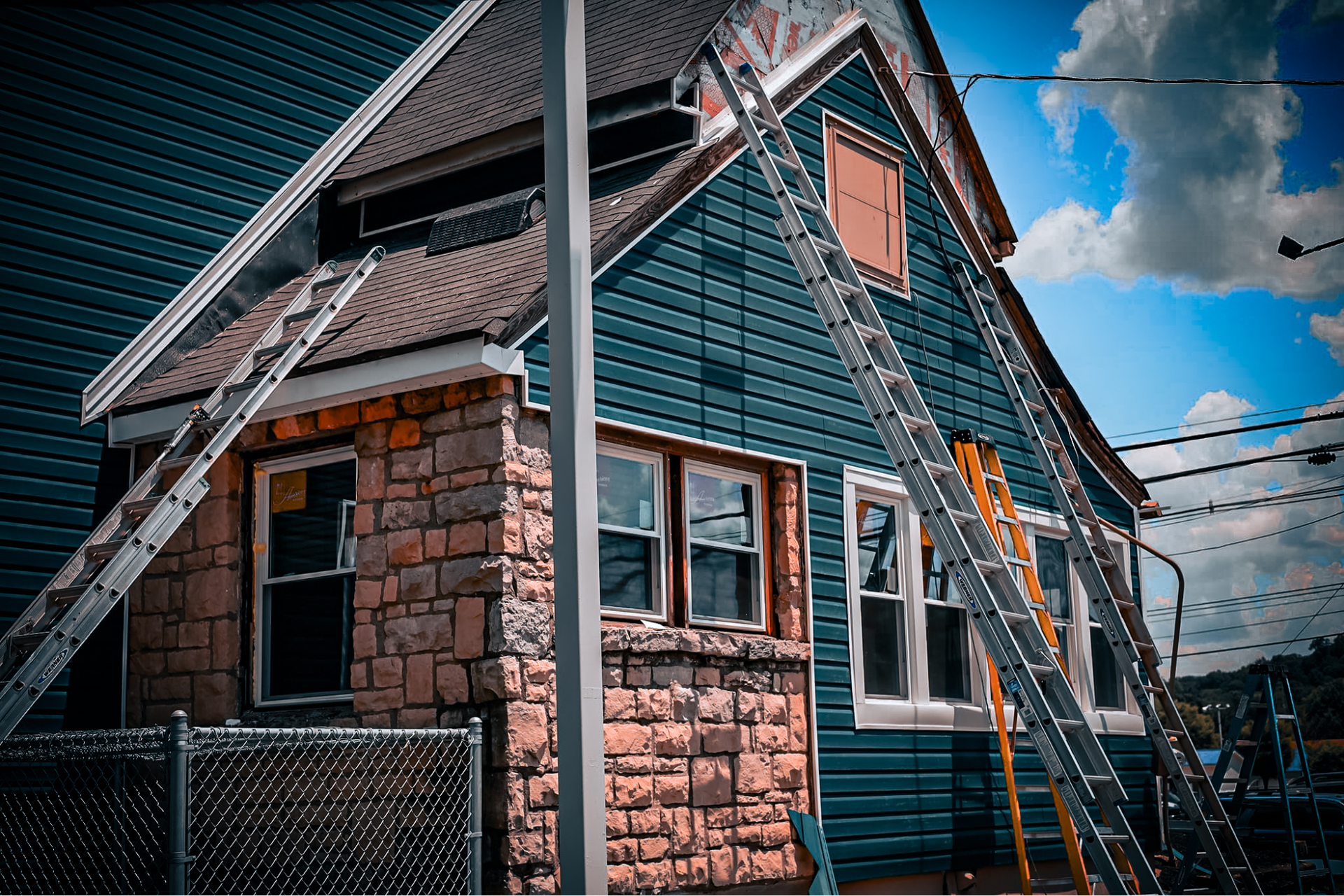 House exterior under construction, blue siding, stone facade, ladders propped against the walls, sunny sky.