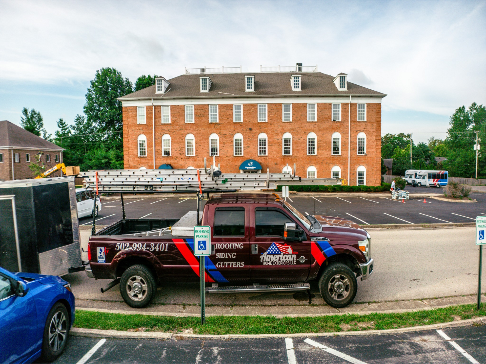 Red truck with ladder on roof in front of brick building, blue car and black trailer in parking lot.