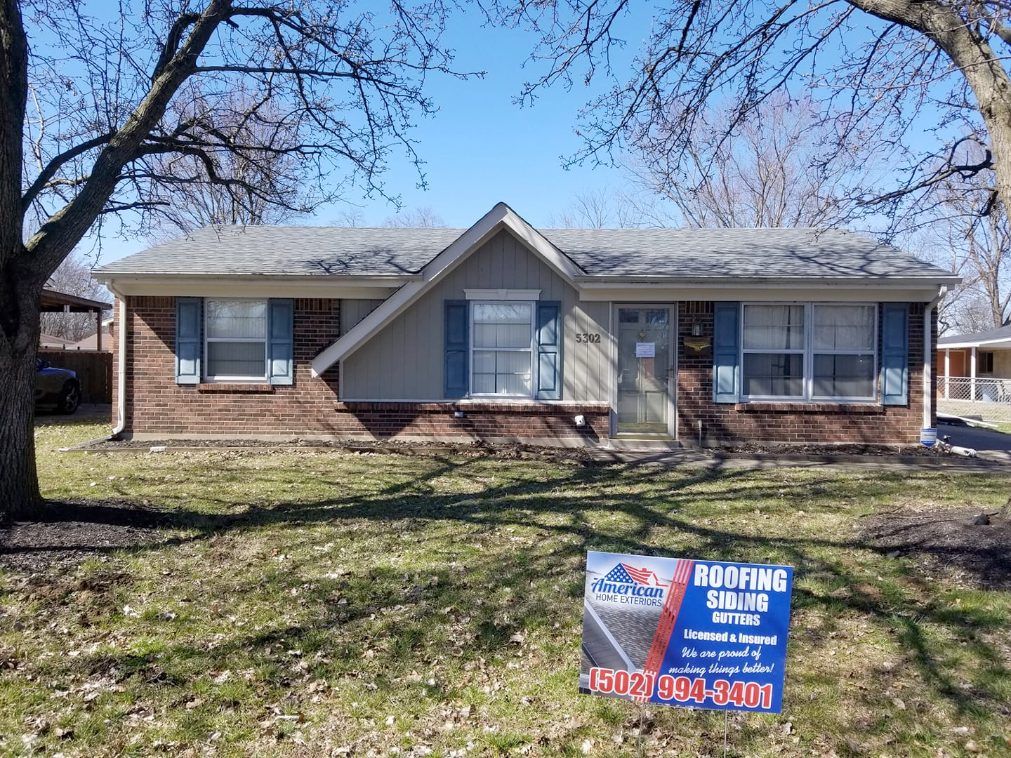 A brick house with blue shutters and a sign in front of it.