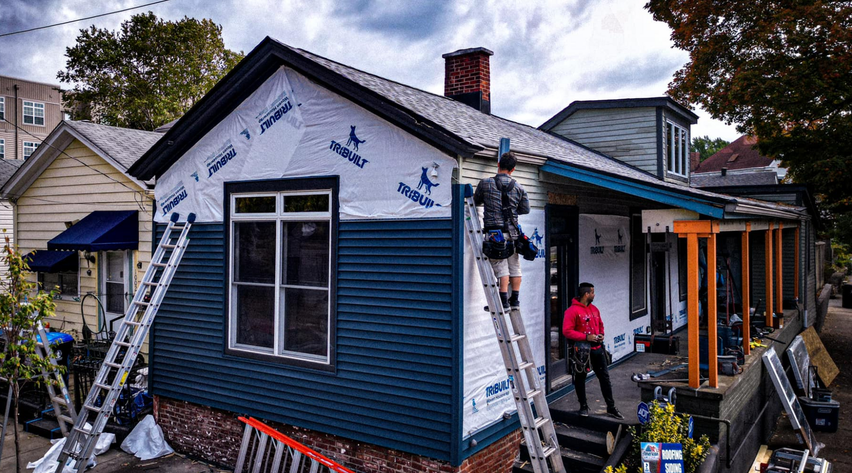 House under construction, with workers on ladders applying siding. Blue siding, white wrap, brick chimney.