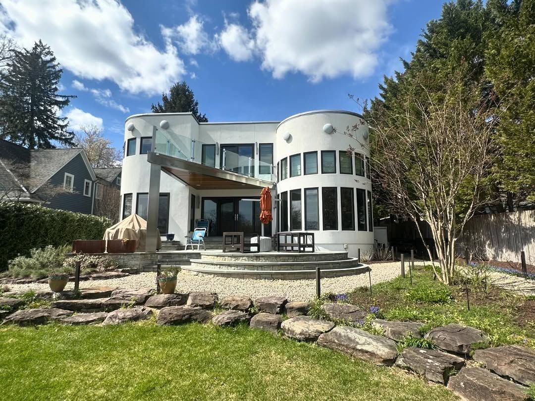 A modern two-story home with rounded white towers, a stone patio, and a landscaped backyard under a sunny, cloudy sky.