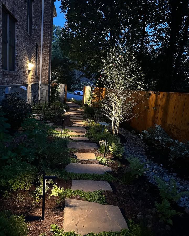 A stone pathway winds along a house exterior at twilight, illuminated by path lights and an uplighted tree.