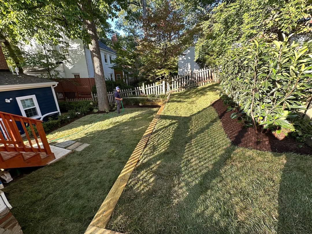A backyard view featuring a blue house with wooden stairs, a fresh green lawn, a wooden border, and a fenced yard perimeter.