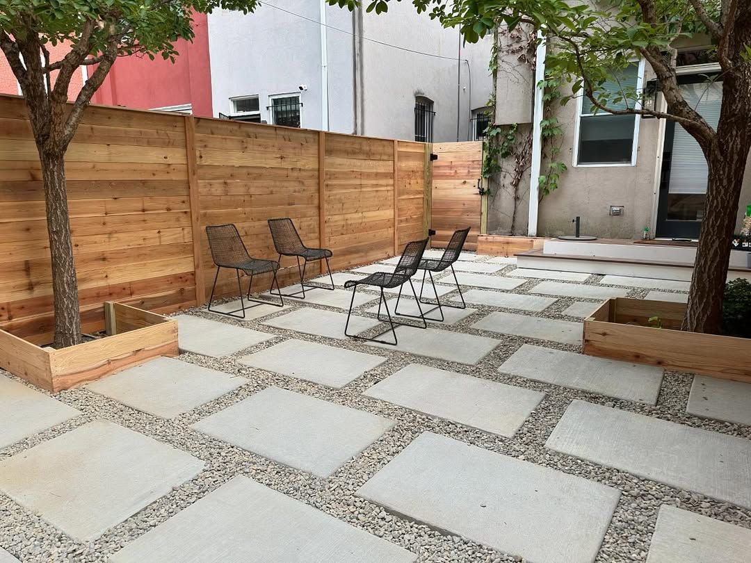 A paved courtyard with square concrete tiles and gravel borders, featuring four metal chairs and two wooden tree planters.
