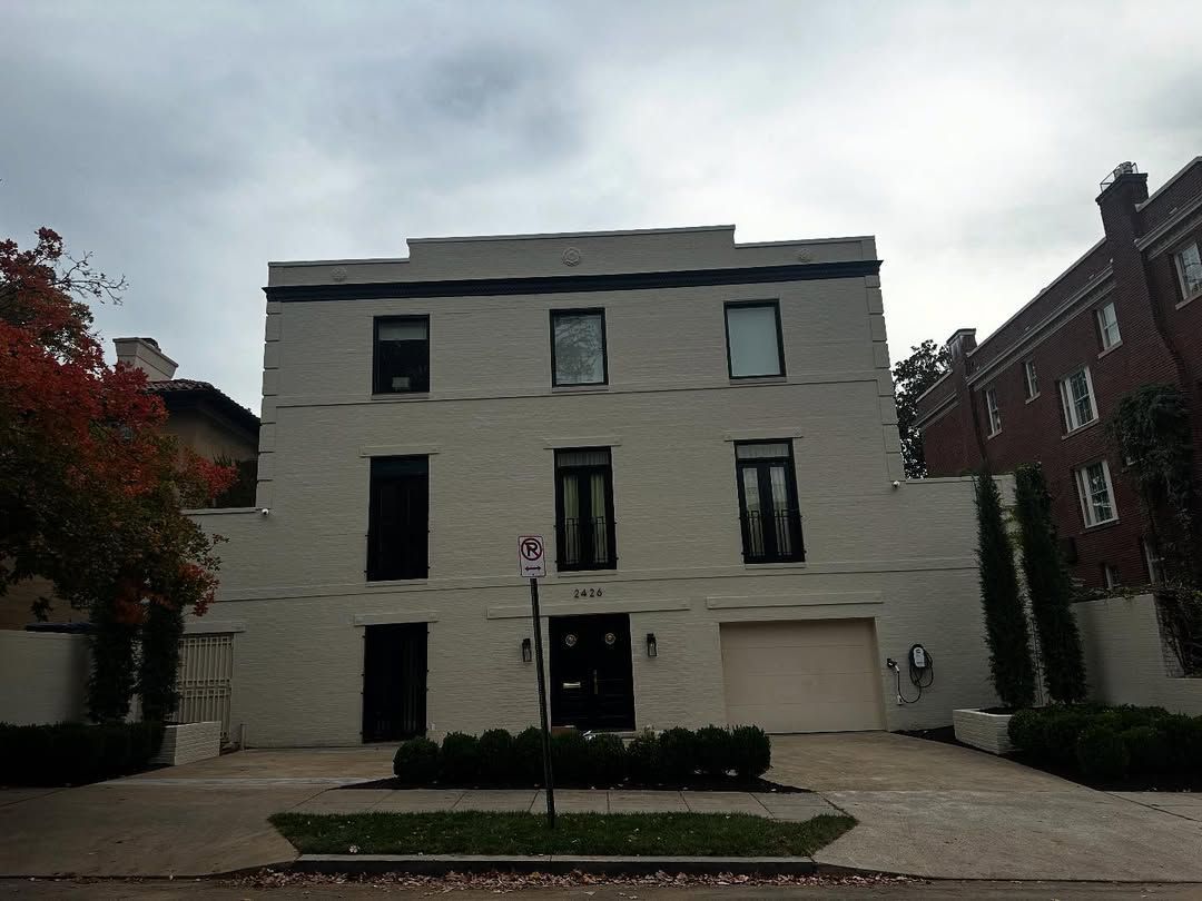 A three-story, light-colored brick house with dark window frames and an attached garage, fronted by a paved driveway.