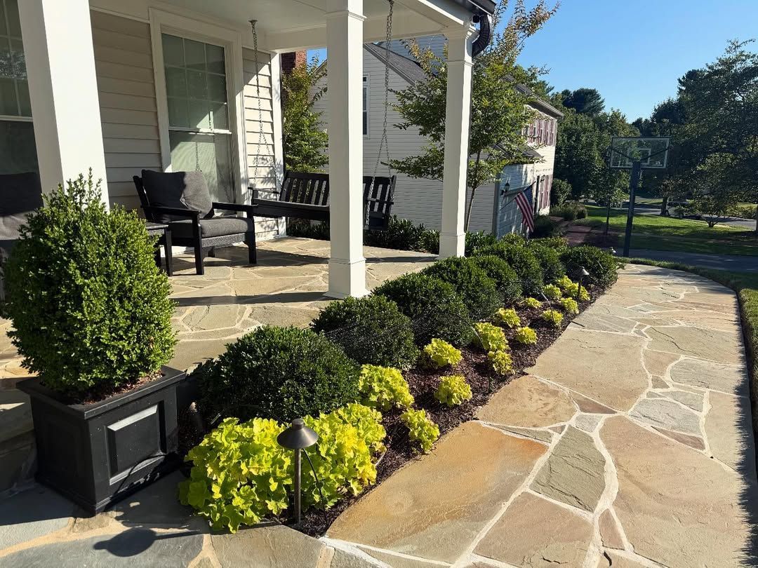 A stone patio with a porch, bench, potted shrub, and a garden border of green shrubs and bright yellow plants.