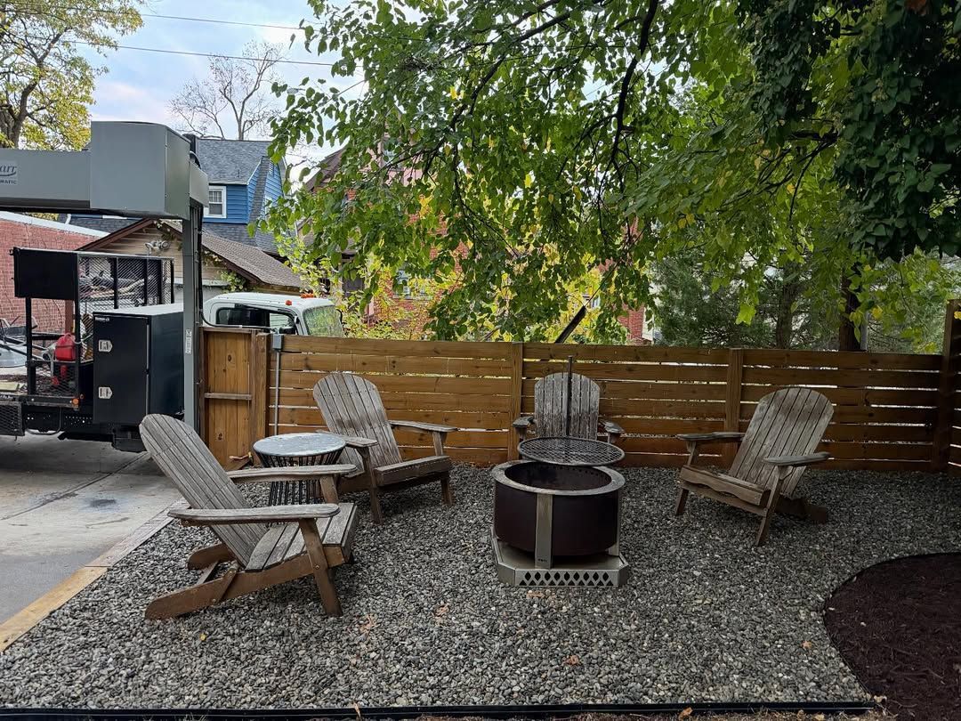 A cozy outdoor patio area featuring a fire pit, four wicker-style chairs, and a small round table on a gravel ground.