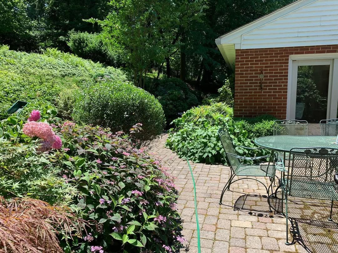 A paved courtyard with square concrete tiles and gravel borders, featuring four metal chairs and two wooden tree planters.