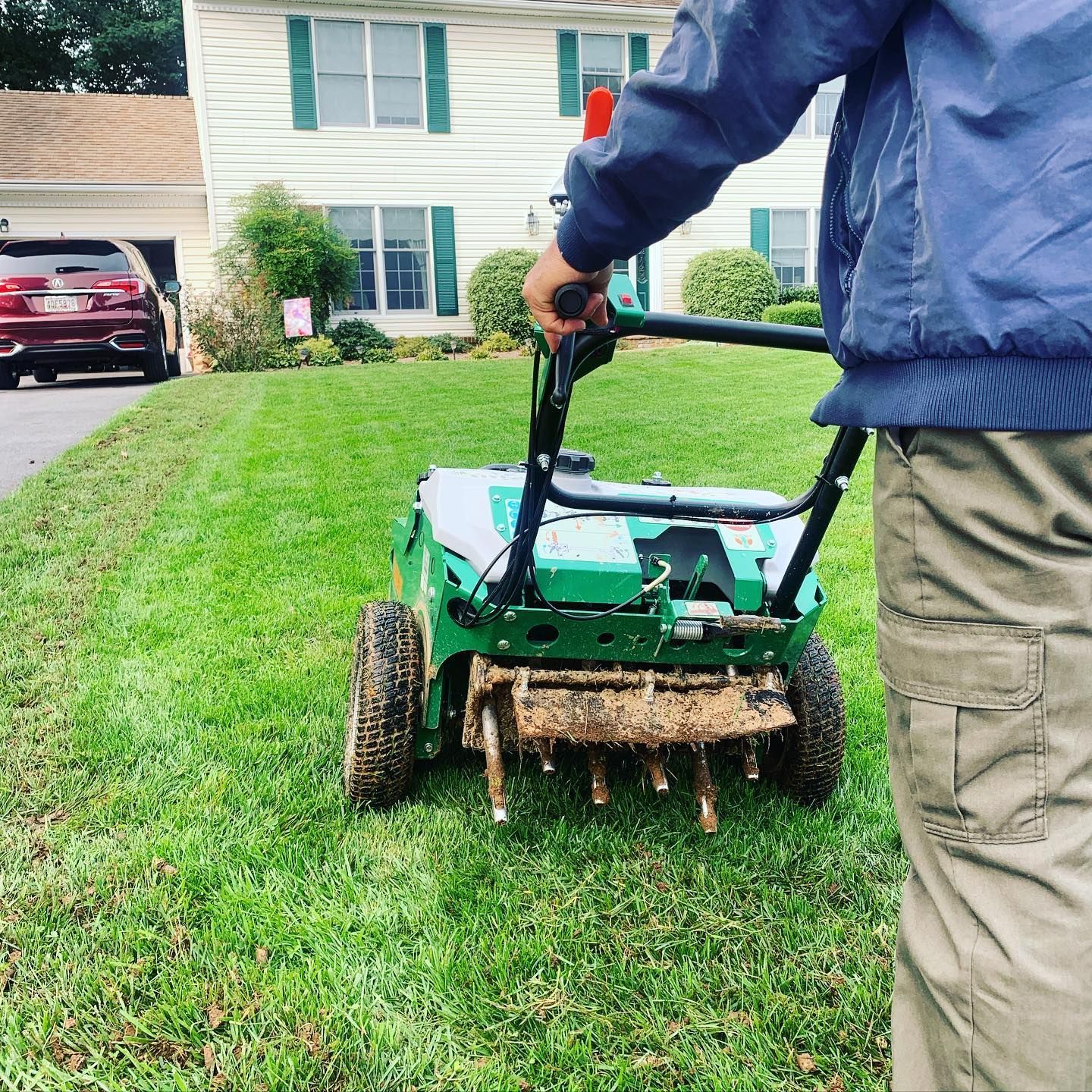 A person in a blue jacket and khaki pants uses a green lawn aerator to pull plugs of soil from a grassy yard.