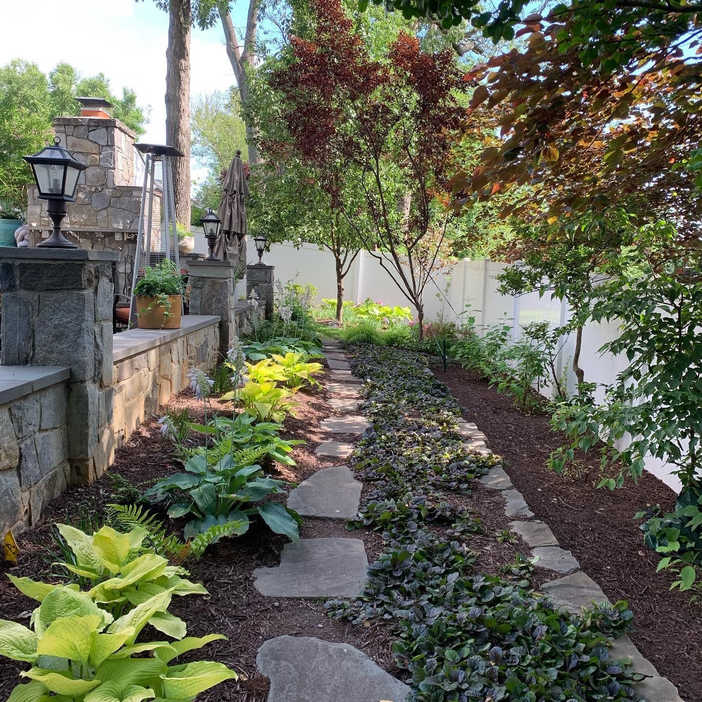 A stone path leads past a house with two AC units and a side garden toward a tree with orange autumn leaves.