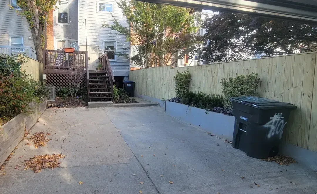 A paved residential backyard with a wooden deck, a tall fence, a raised planter box, and a black trash bin.