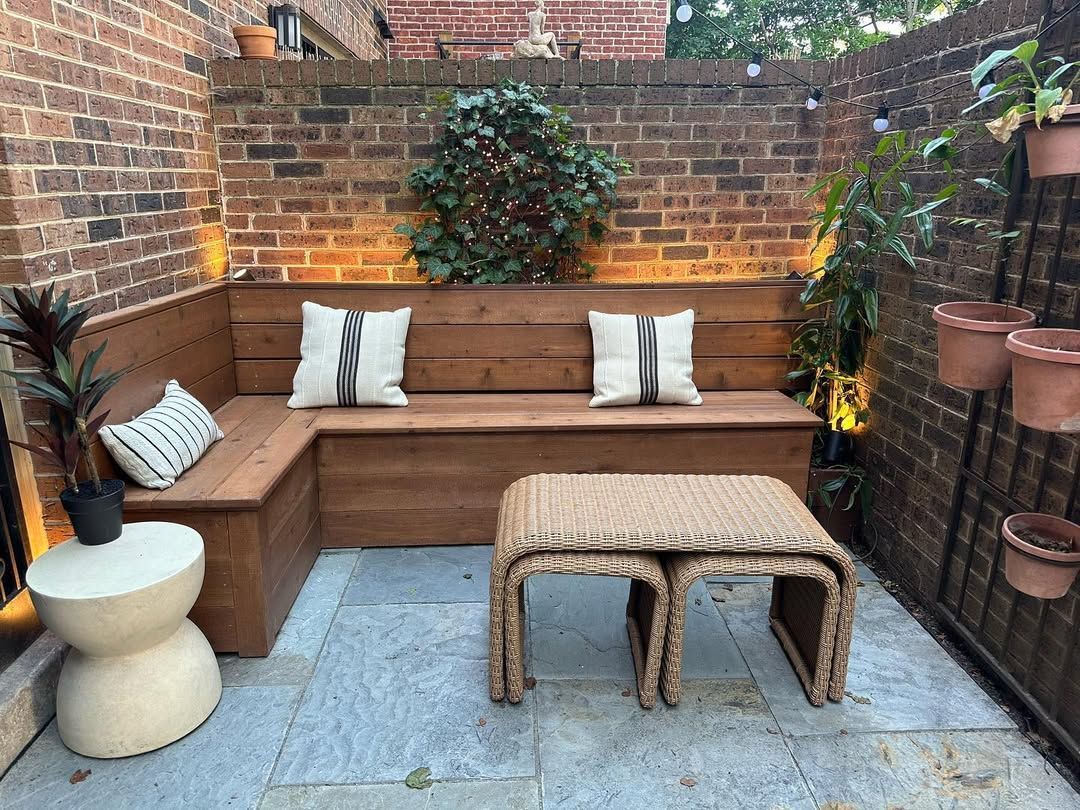 An L-shaped wooden bench with white throw pillows, a woven coffee table, and potted plants in a brick-walled patio.