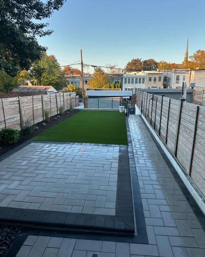 A modern backyard with a rectangular lawn, grey herringbone-patterned stone patio, and high wooden privacy fences.