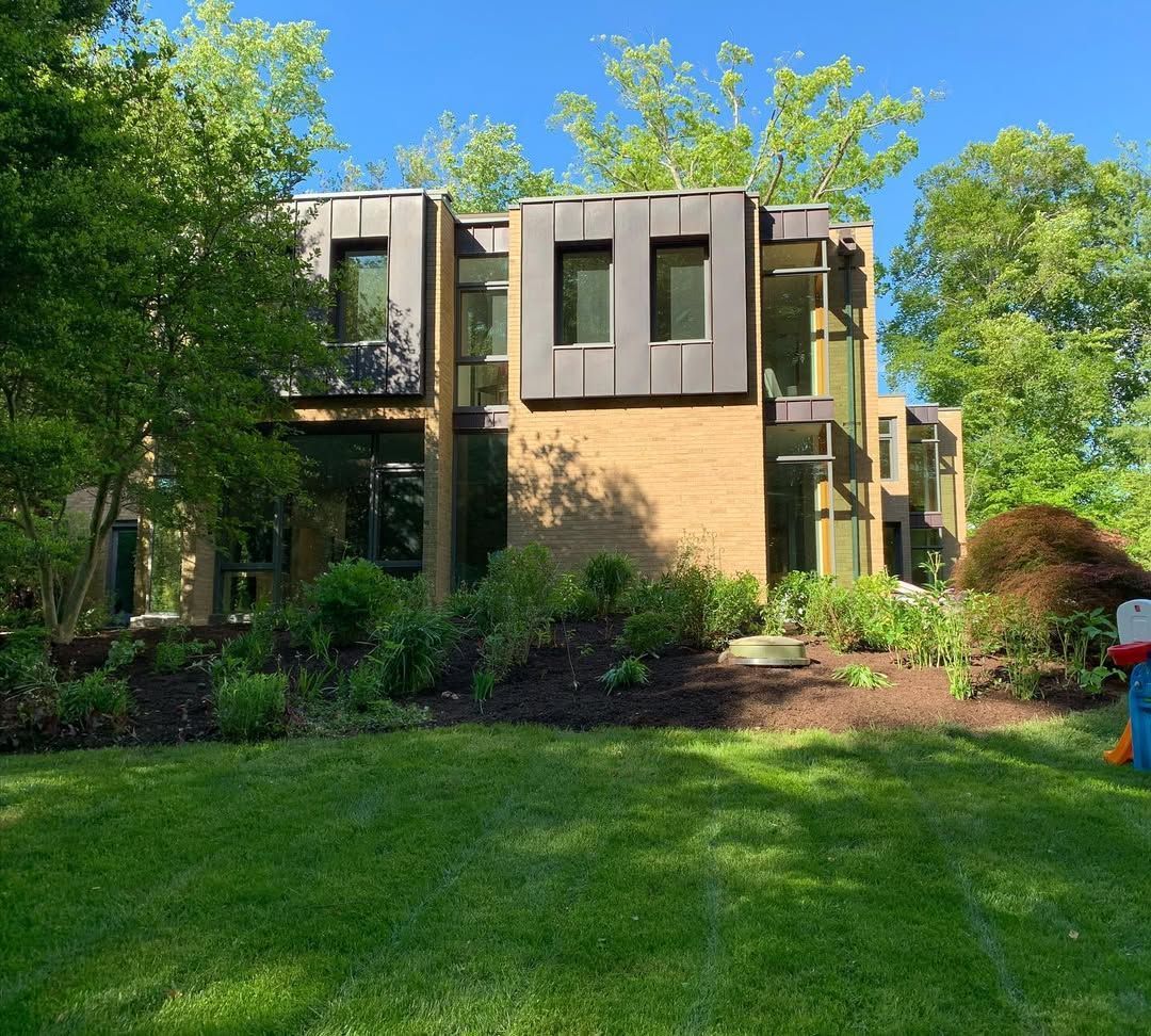 A two-story modern house with tan brick, dark panels, and large windows, set behind a lush green lawn and trees.