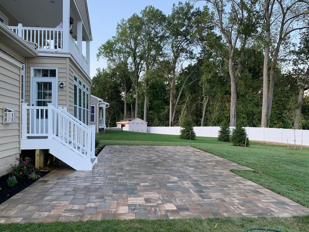 A tan two-story house with a white staircase leading to a new stone paver patio in a grassy backyard with a white fence.