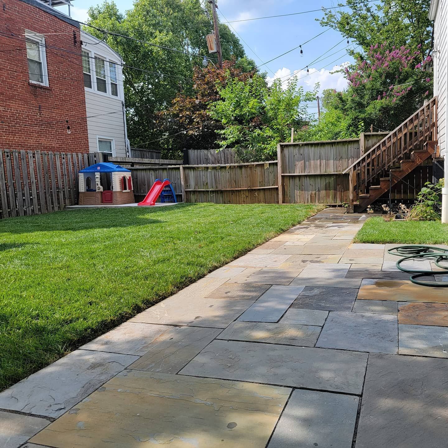 A backyard with a stone patio in the foreground, a grassy lawn, a small playhouse, and a wooden staircase to a building.