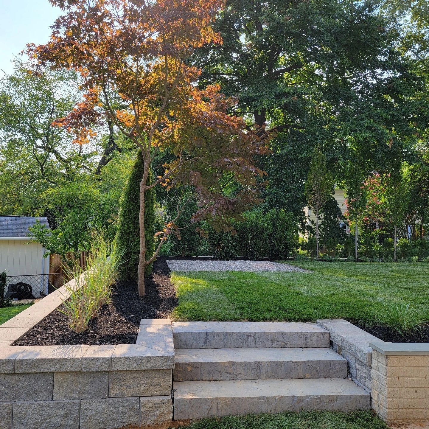 Stone steps lead up to a tiered backyard featuring a mulch garden bed with a small tree and a patch of green grass.