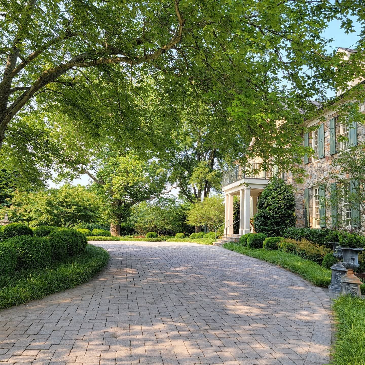 A curved brick driveway leads toward a light-colored multi-story home surrounded by lush green trees and landscaping.