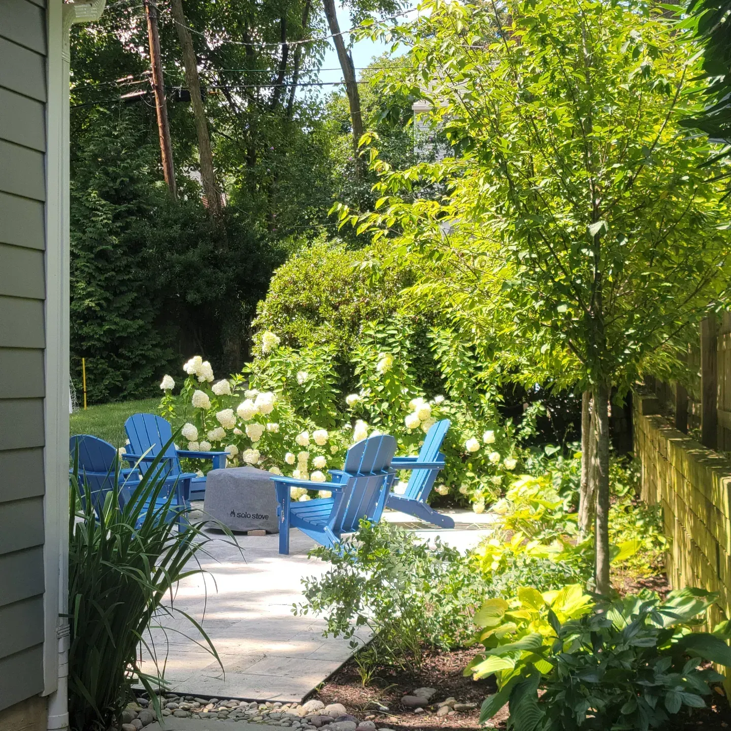 Blue Adirondack chairs arranged around a small fire pit on a stone patio in a lush, green, sunlit backyard garden.