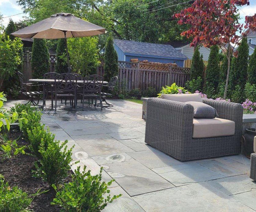A stone patio in a backyard features a dining table with an umbrella, wicker lounge chairs, and manicured green hedges.