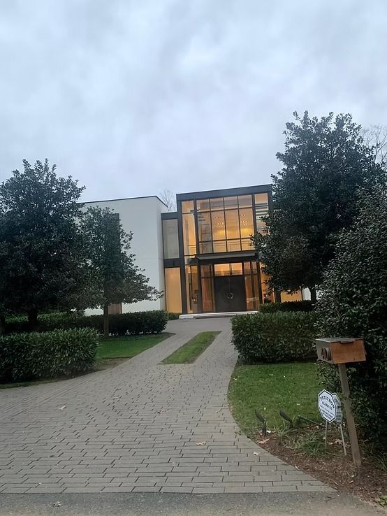 A modern two-story building with a large glass-paneled entrance and a white exterior, surrounded by trees and shrubs.