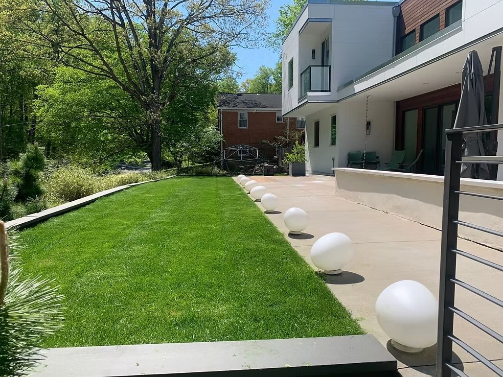 A green lawn runs alongside a patio featuring a row of spherical outdoor lights near a house wall.