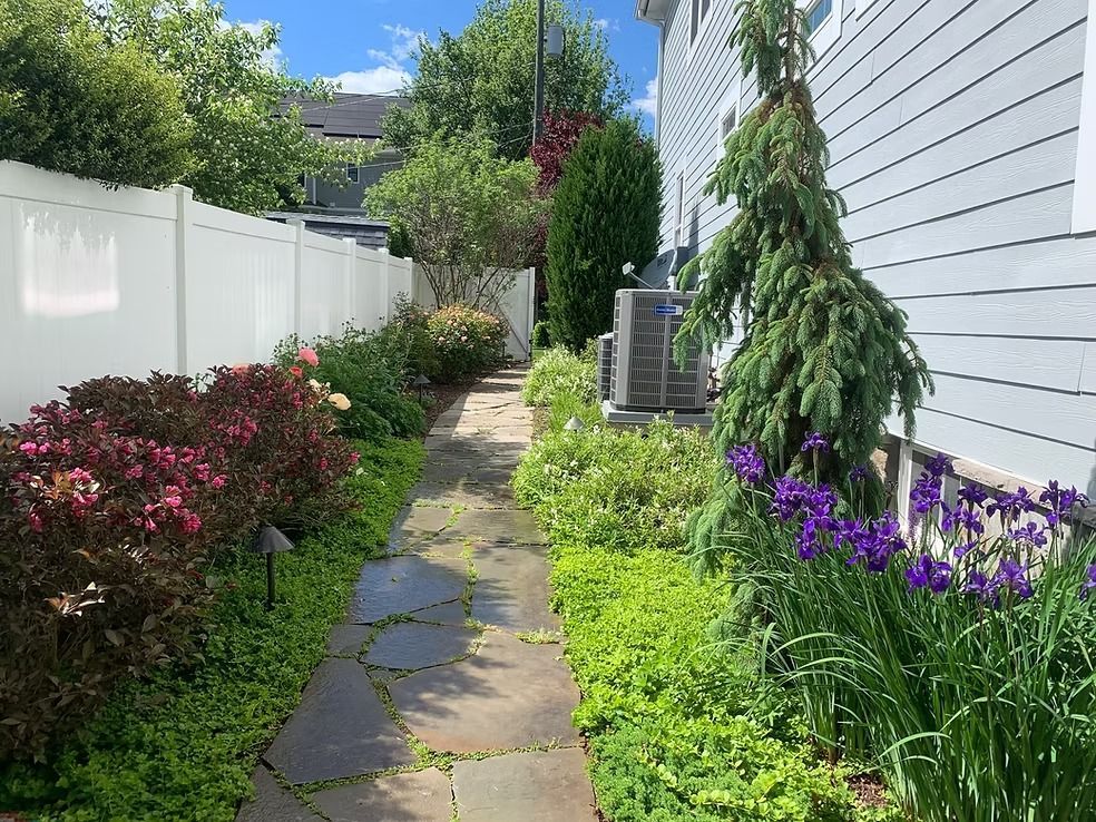 A stone path winds through a lush backyard garden beside a house, bordered by colorful flowers, shrubs, and a white fence.