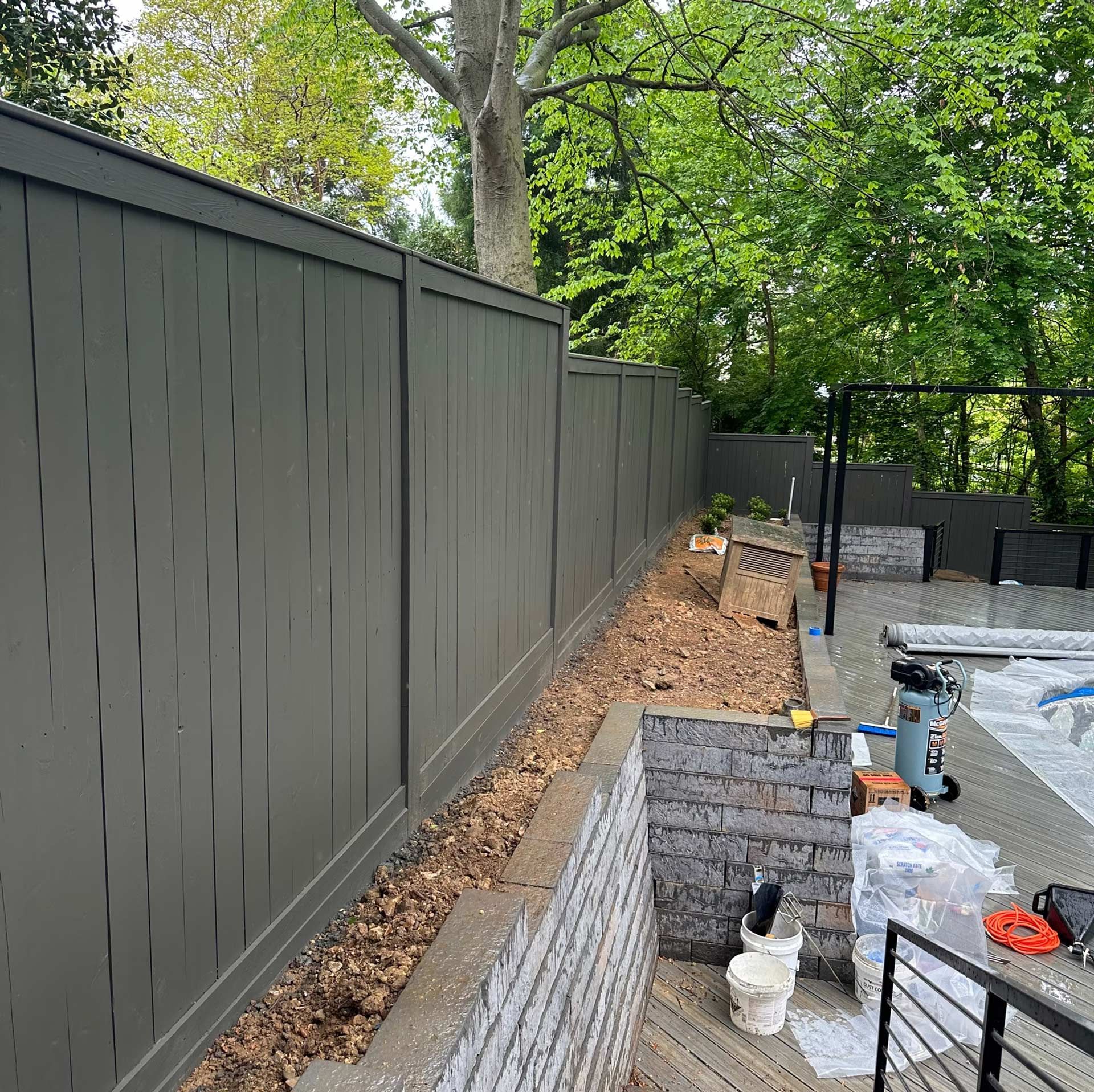 A grey privacy fence sits behind a tiered stone retaining wall in a backyard with trees and a pool deck area.