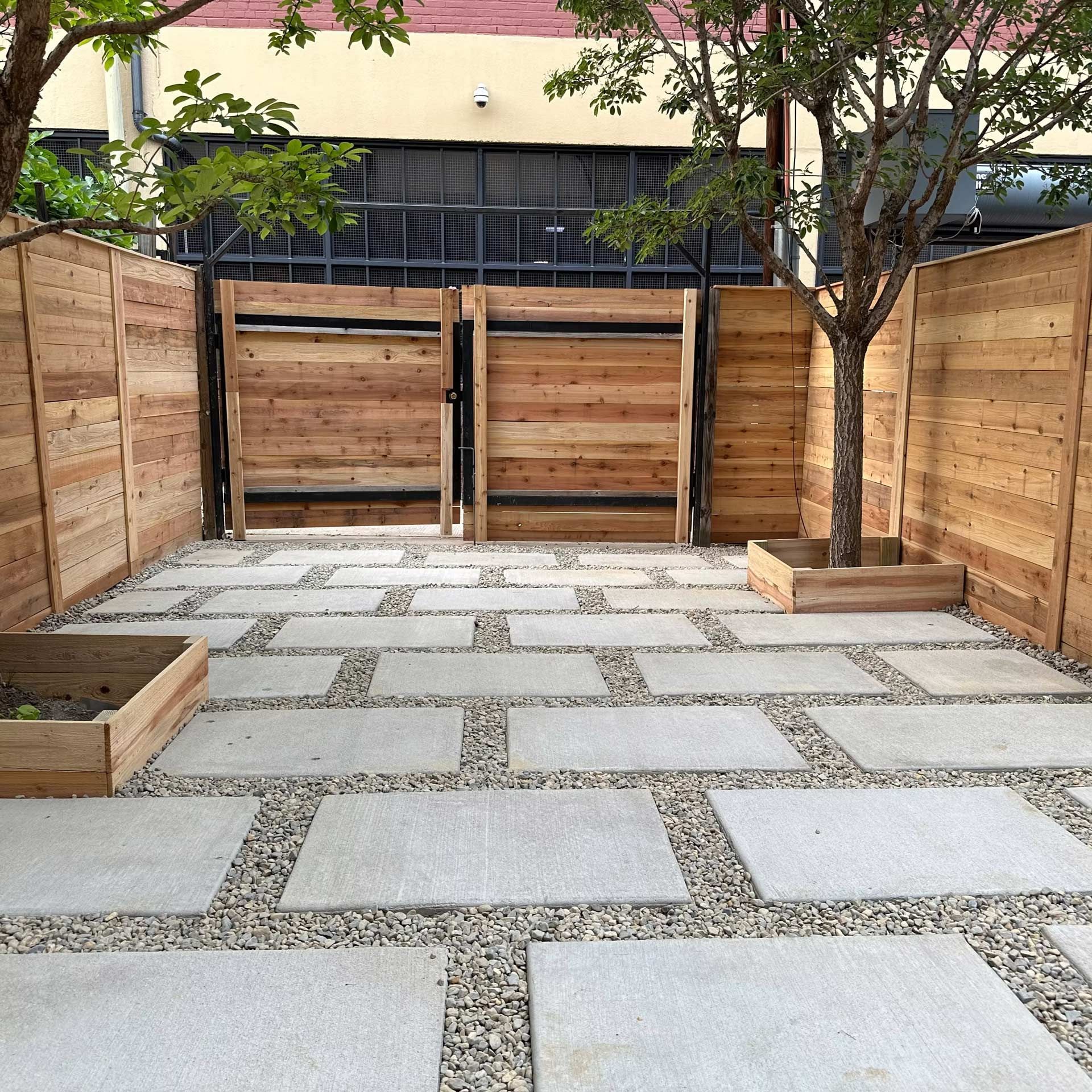 A modern patio with light grey square pavers separated by gravel, framed by wooden fencing, planters, and a small tree.