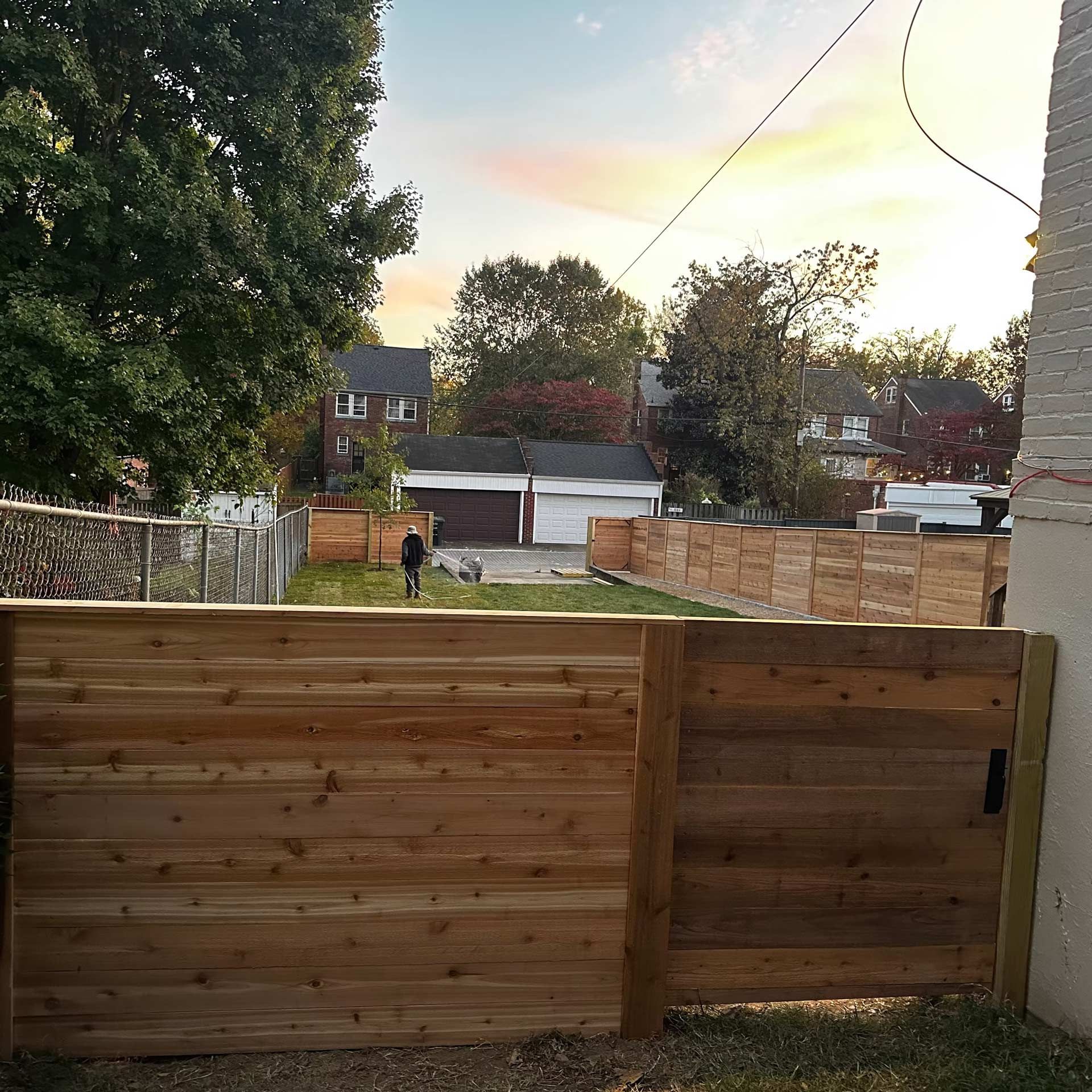 A horizontal wooden privacy fence stands in the foreground of a yard, with a person standing in the grass beyond it.