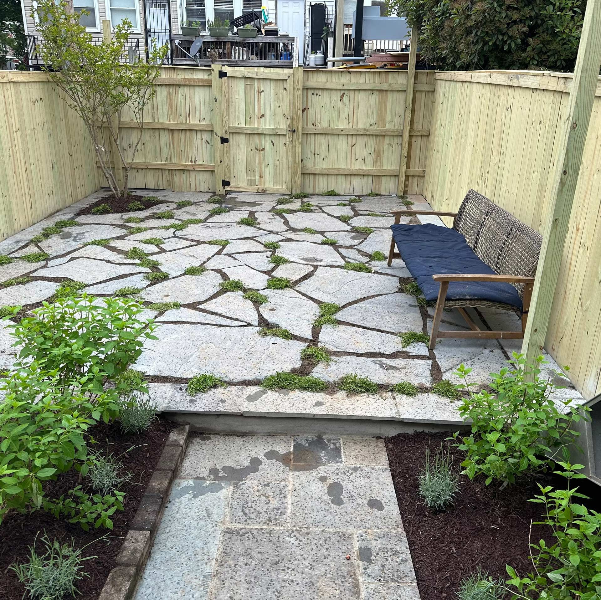 A stone patio with a blue bench inside a small wooden fenced yard with garden beds in the foreground.