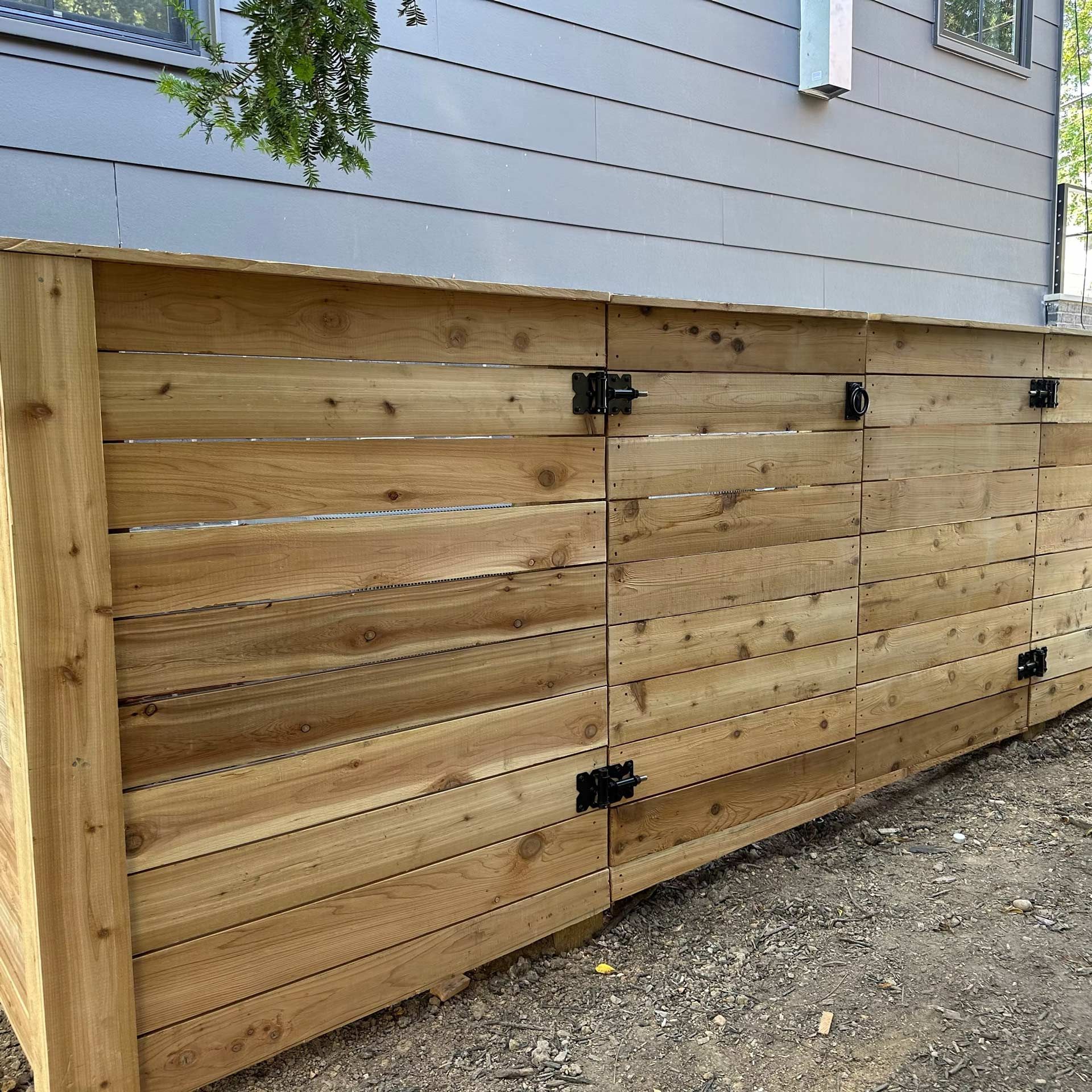 A wooden horizontal slat privacy screen for garbage cans against a house exterior wall.