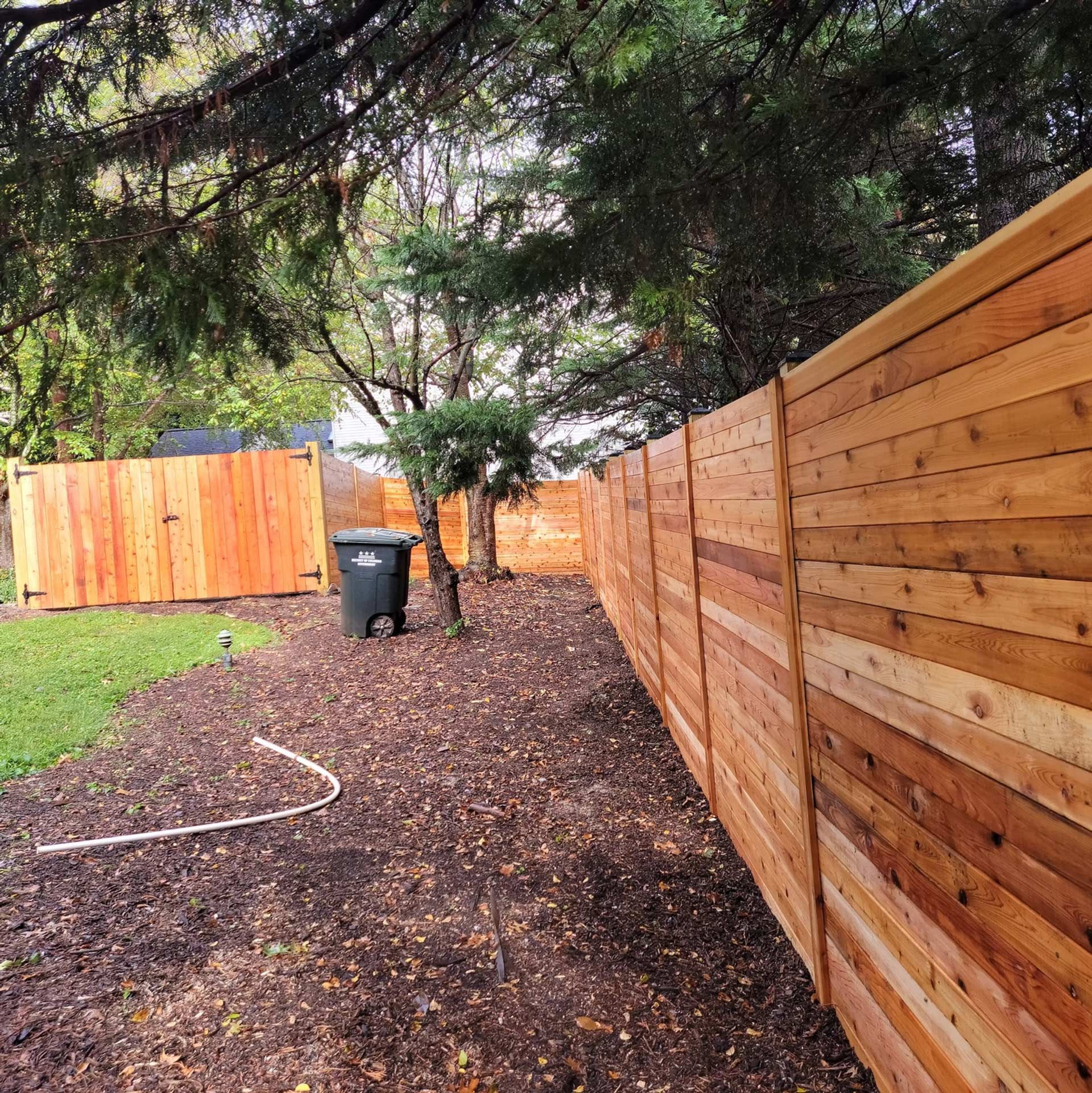 A newly installed horizontal cedar wood fence runs through a backyard with mulch and a trash bin near the corner.