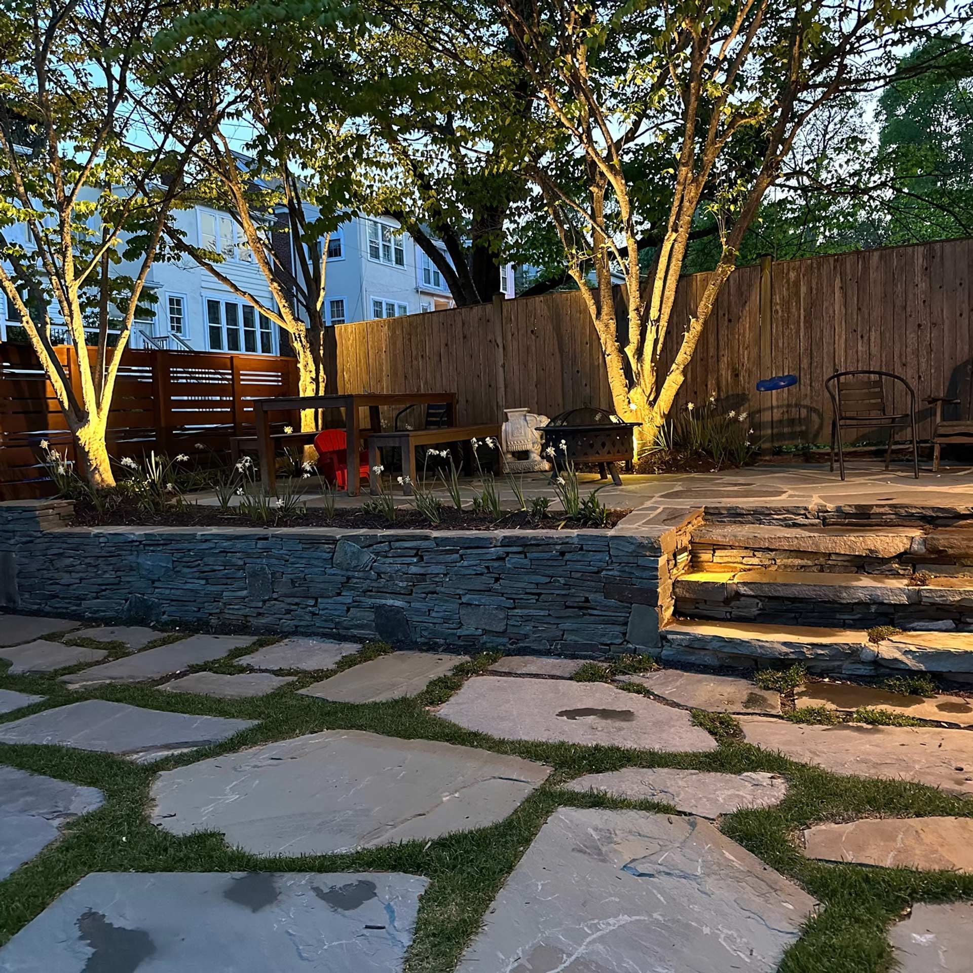 A flagstone patio leads to a stone retaining wall and steps, with outdoor seating areas beneath trees and a wooden fence.